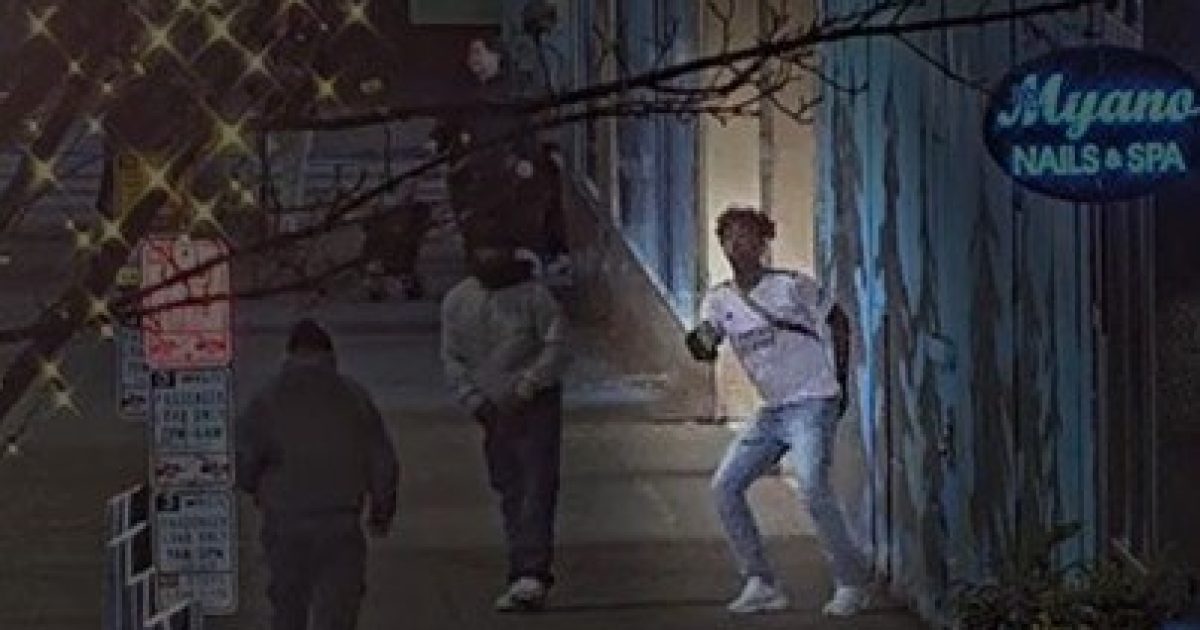Three individuals on a city sidewalk at night, with one person appearing to hold an object, near a nail salon sign.