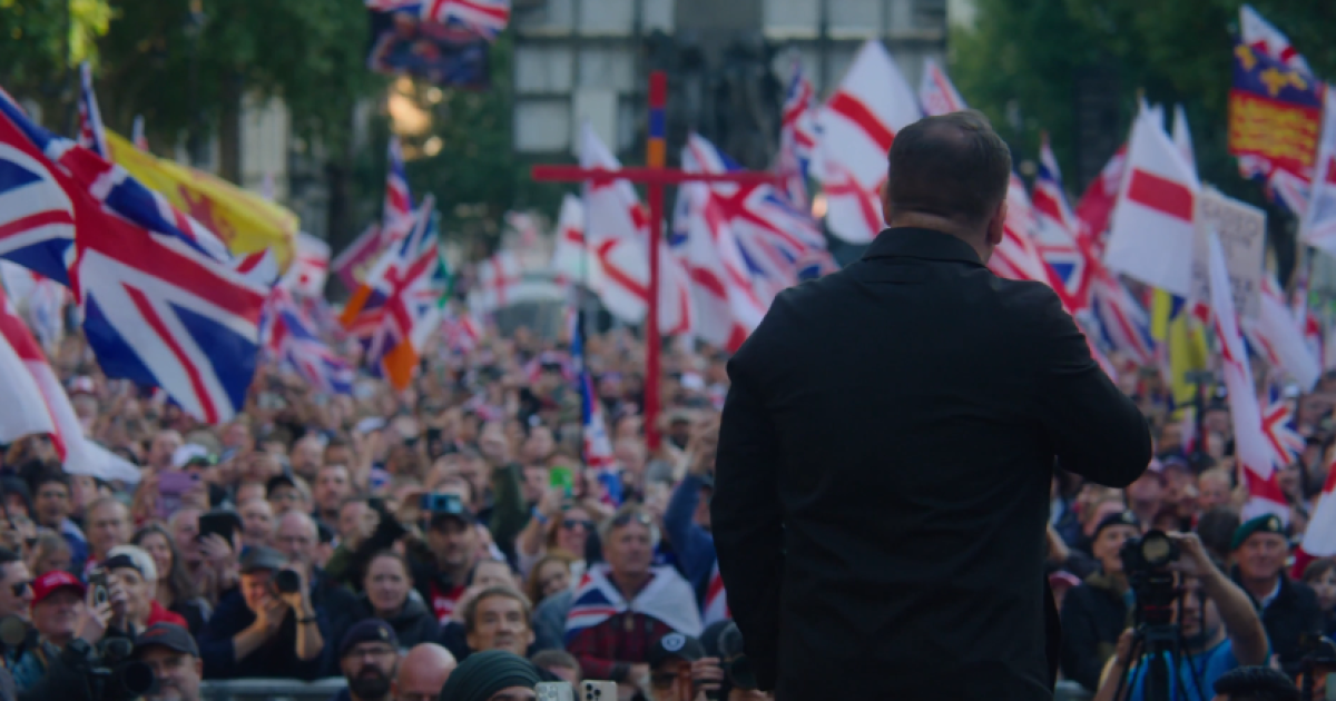 Crowd of supporters waving British flags at a political rally, with a speaker addressing the audience from the stage.
