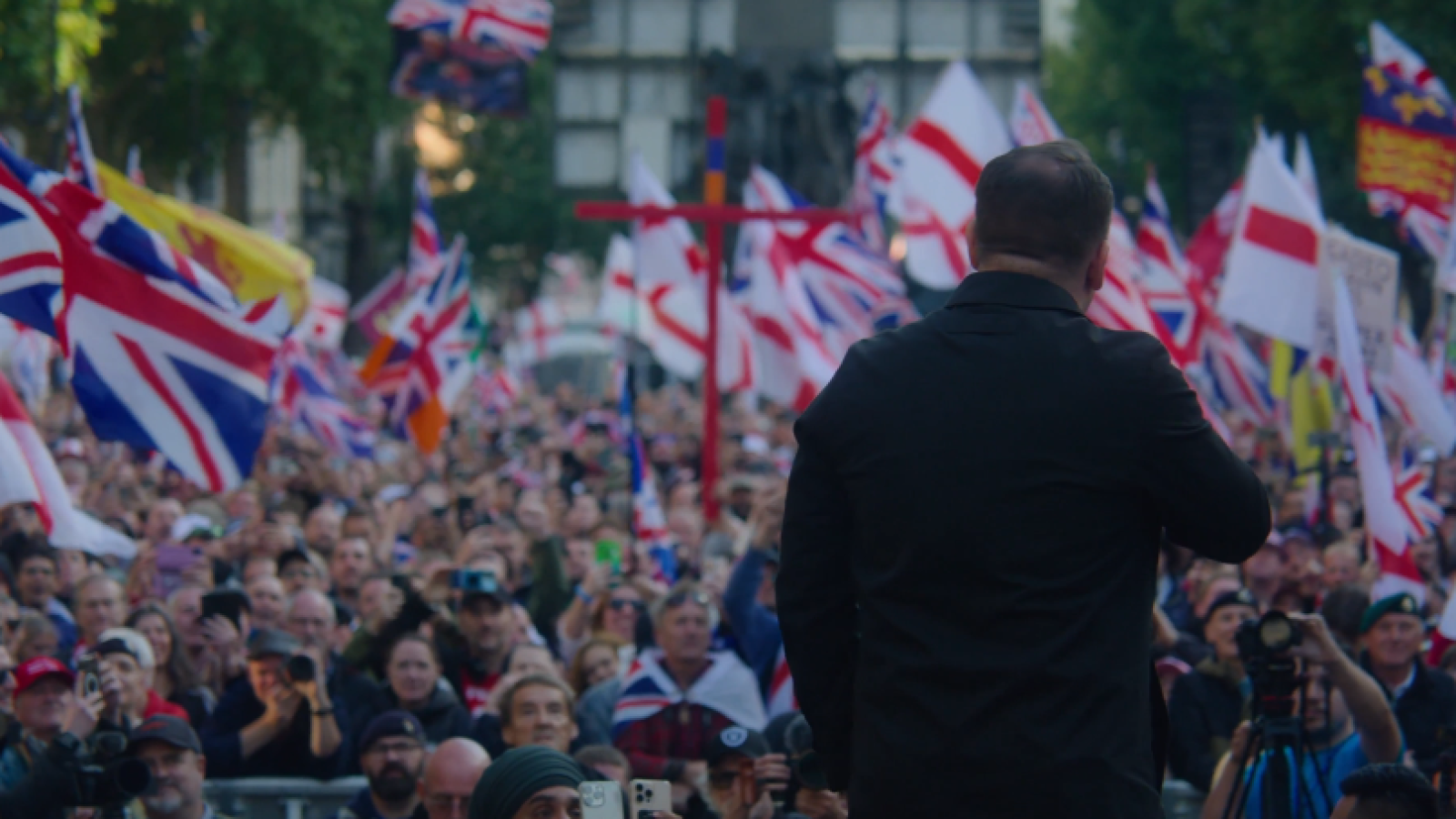 Crowd of supporters waving British flags at a political rally, with a speaker addressing the audience from the stage.