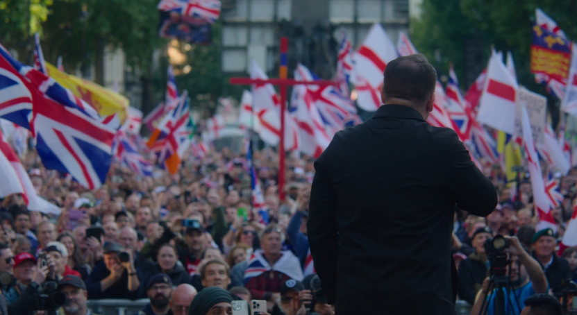 Crowd of supporters waving British flags at a political rally, with a speaker addressing the audience from the stage.