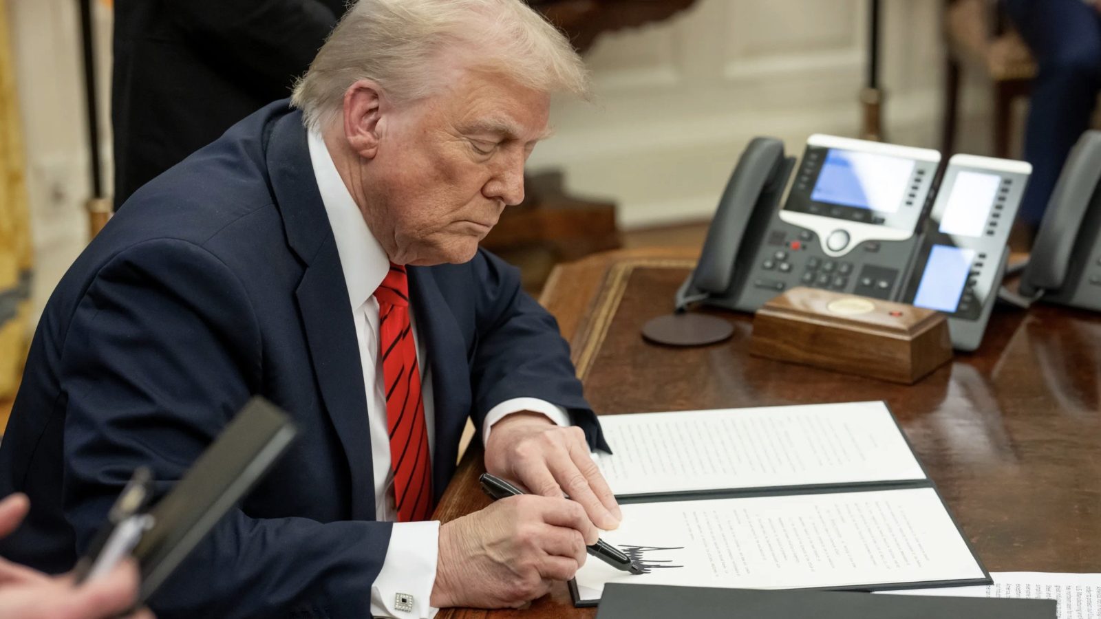 Donald Trump signs a document at a desk in the Oval Office, surrounded by office equipment and staff members.