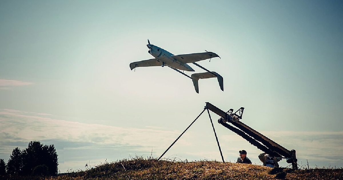 A drone takes flight from a launch platform while two operators observe the launch in an outdoor setting.