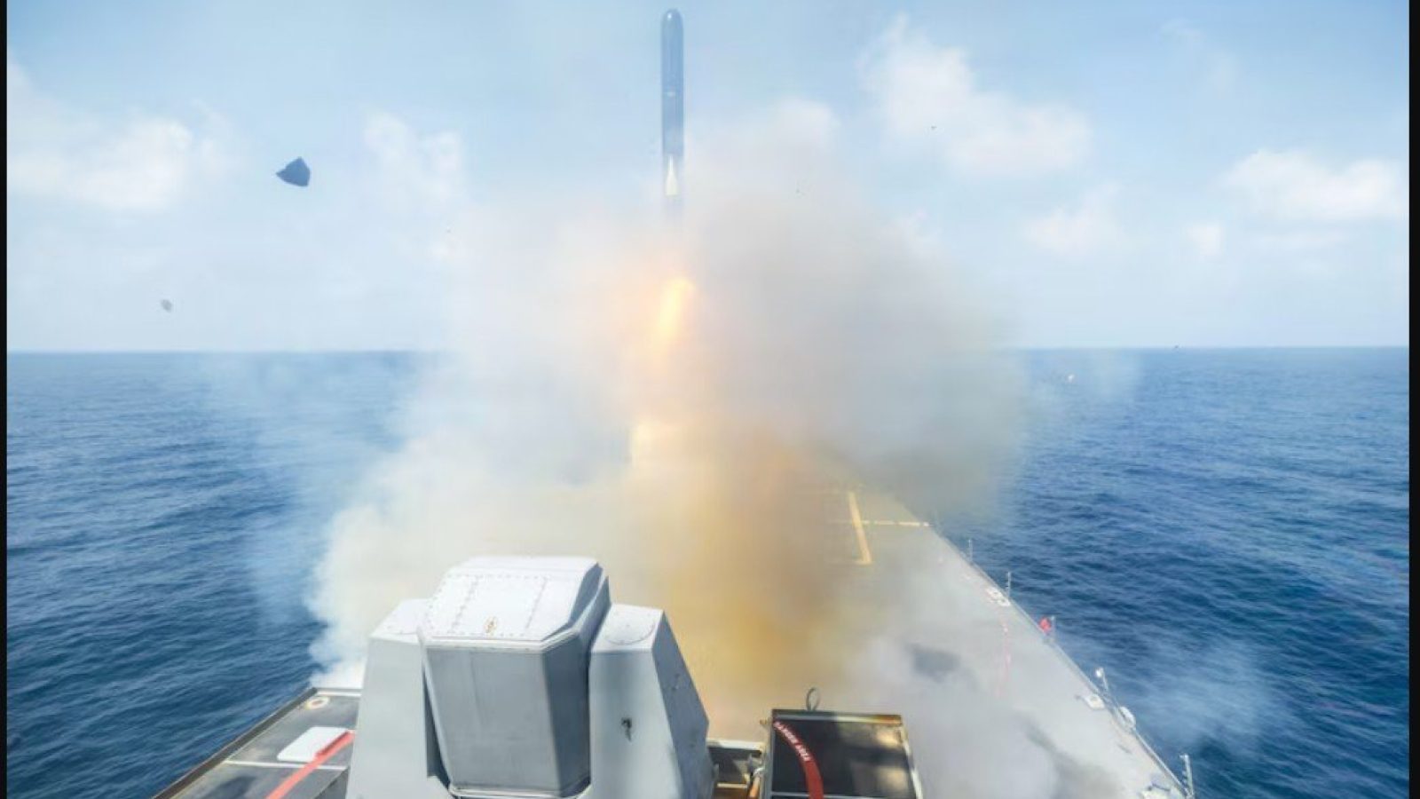 Missile launch from a naval vessel over the ocean, creating smoke and water spray against a clear blue sky.