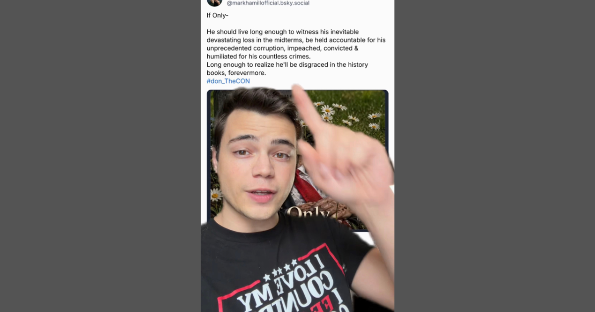 Person gesturing while discussing political accountability, wearing a shirt with a patriotic message, surrounded by flowers in a casual setting.