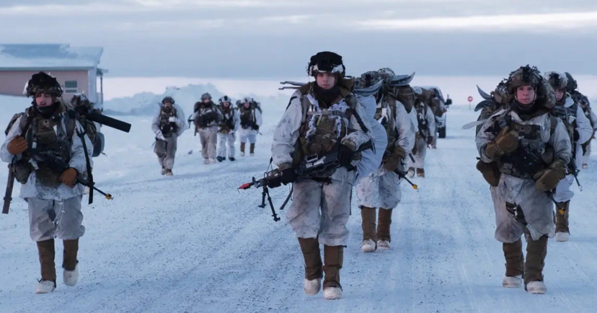 Soldiers in winter camouflage gear march along a snow-covered road, preparing for Arctic training exercises in a cold environment.