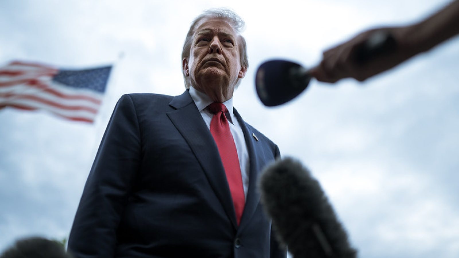 Former President Donald Trump stands before a microphone with an American flag in the background, conveying a serious demeanor during a press event.