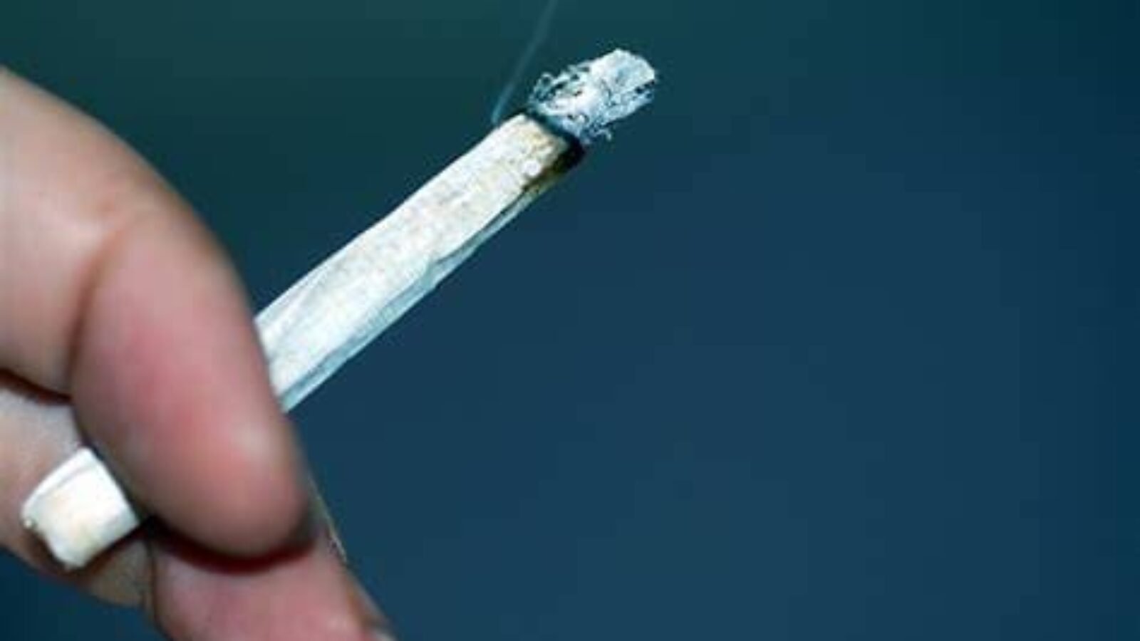 A close-up of a hand holding a burning cigarette with smoke rising against a blurred background.