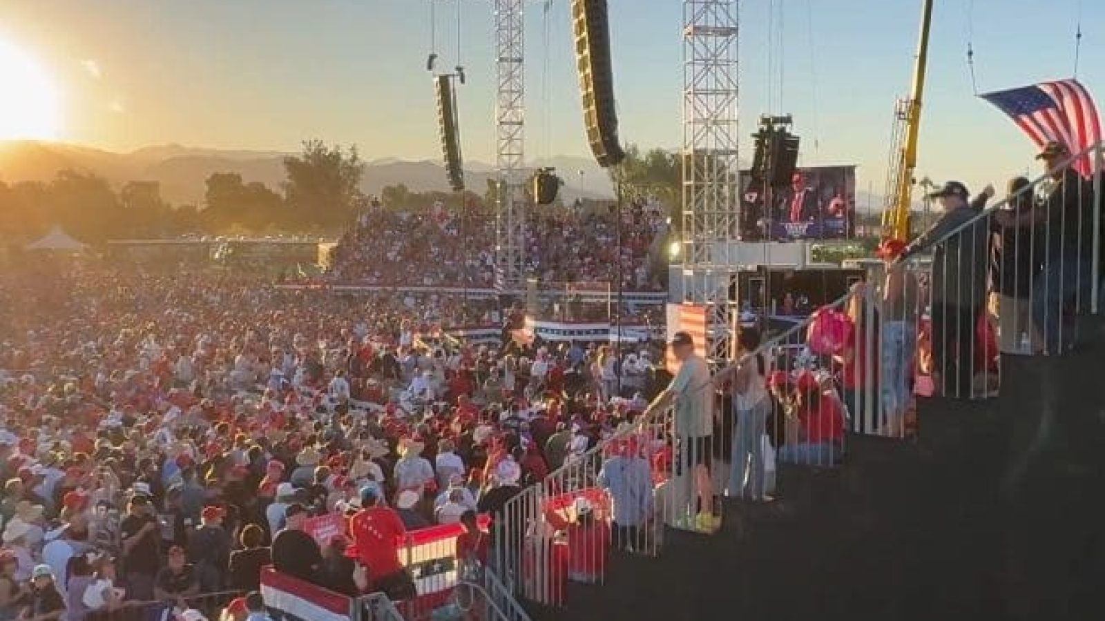 Large crowd attending a political rally at sunset, featuring a stage with speakers and American flags, showcasing a vibrant atmosphere and enthusiastic supporters.