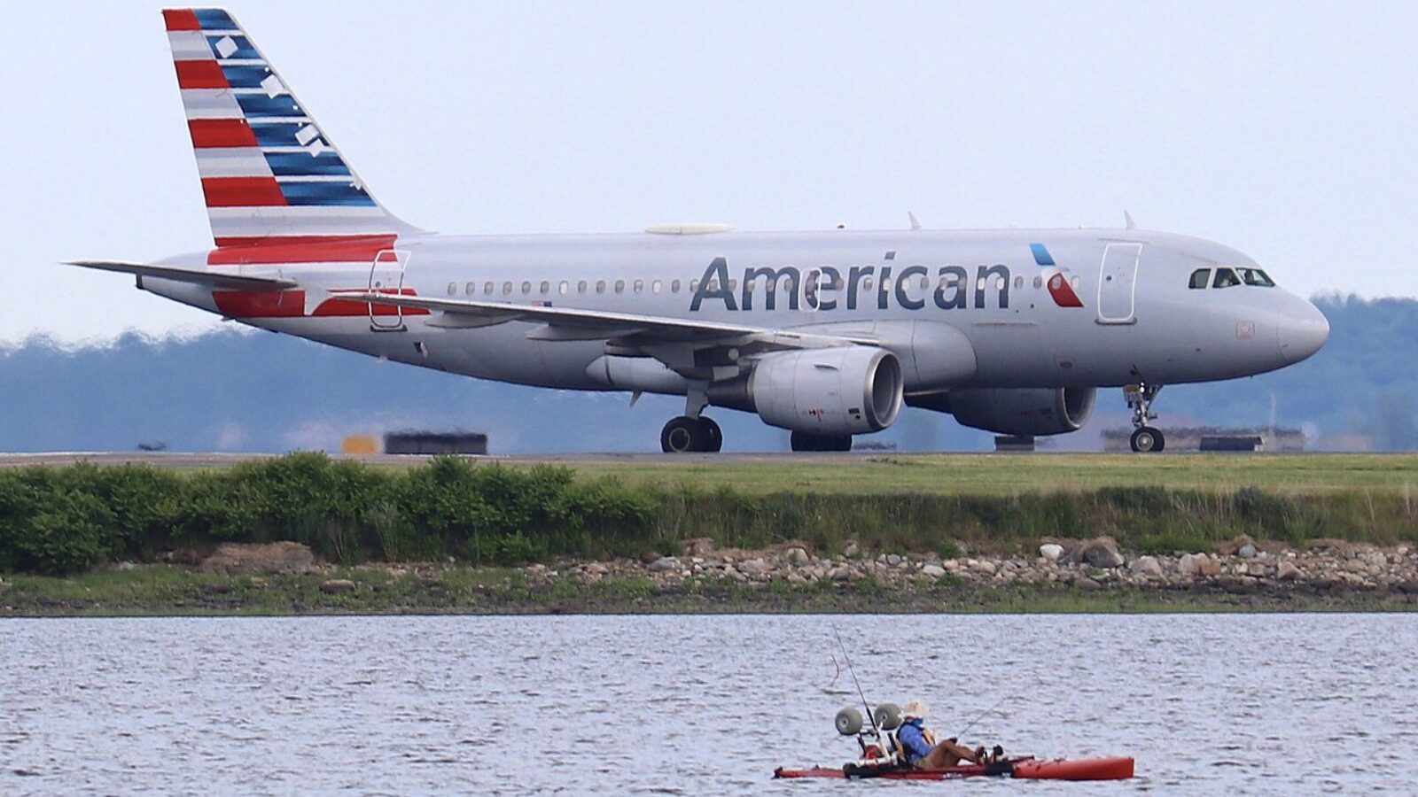 American Airlines plane taxiing near a kayak with two fishermen on a calm waterway.