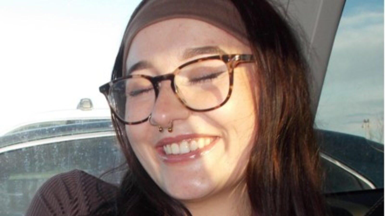 Young woman with glasses and a septum piercing smiling joyfully in a car, showcasing a sunny day and a relaxed atmosphere.