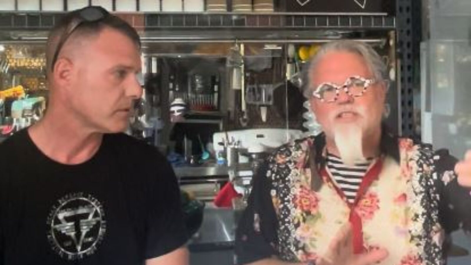 Two men engaged in conversation at a bar, one wearing a floral shirt and glasses, discussing cocktails and mixology techniques.