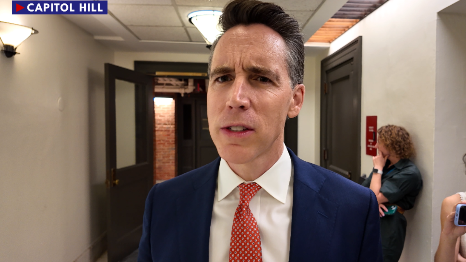 A man in a suit speaks seriously in a hallway on Capitol Hill, with visible doors and lighting fixtures in the background.