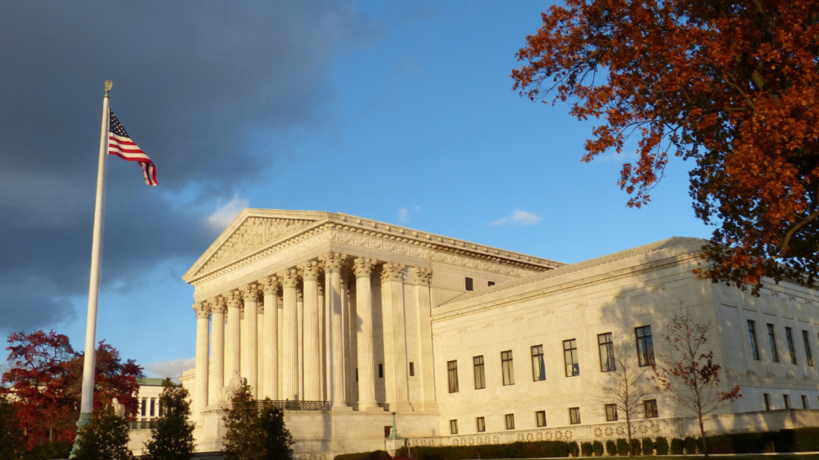 U.S. Supreme Court building with American flag, surrounded by autumn trees and a clear blue sky.