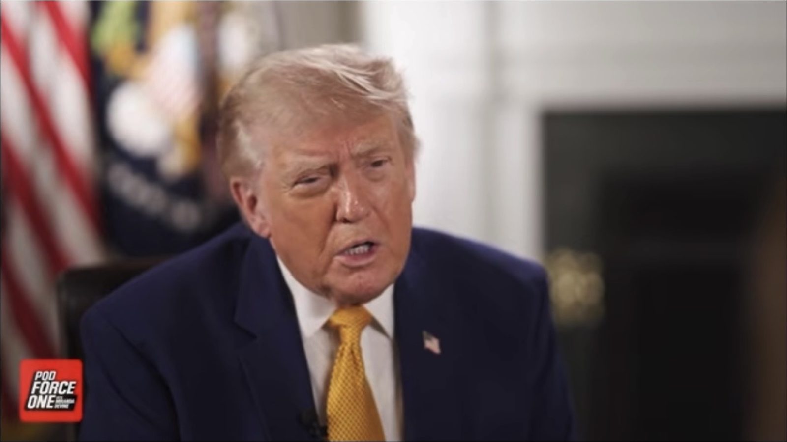 Donald Trump speaking during an interview, wearing a blue suit and yellow tie, with flags and a presidential seal in the background.