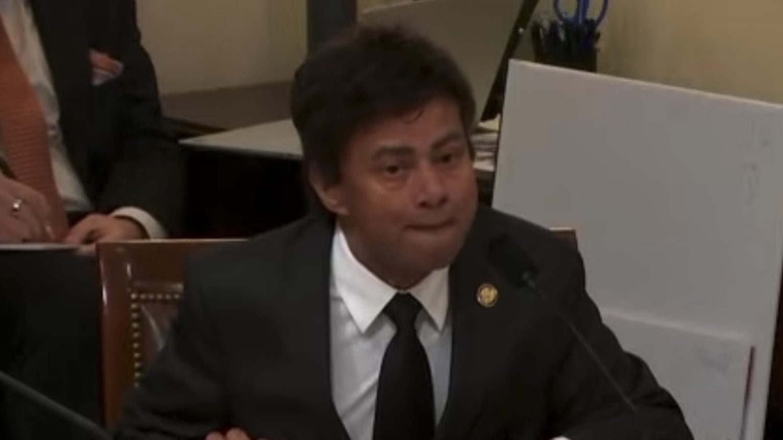 Man in a black suit testifying at a formal hearing, displaying a serious expression, with officials and documents visible in the background.