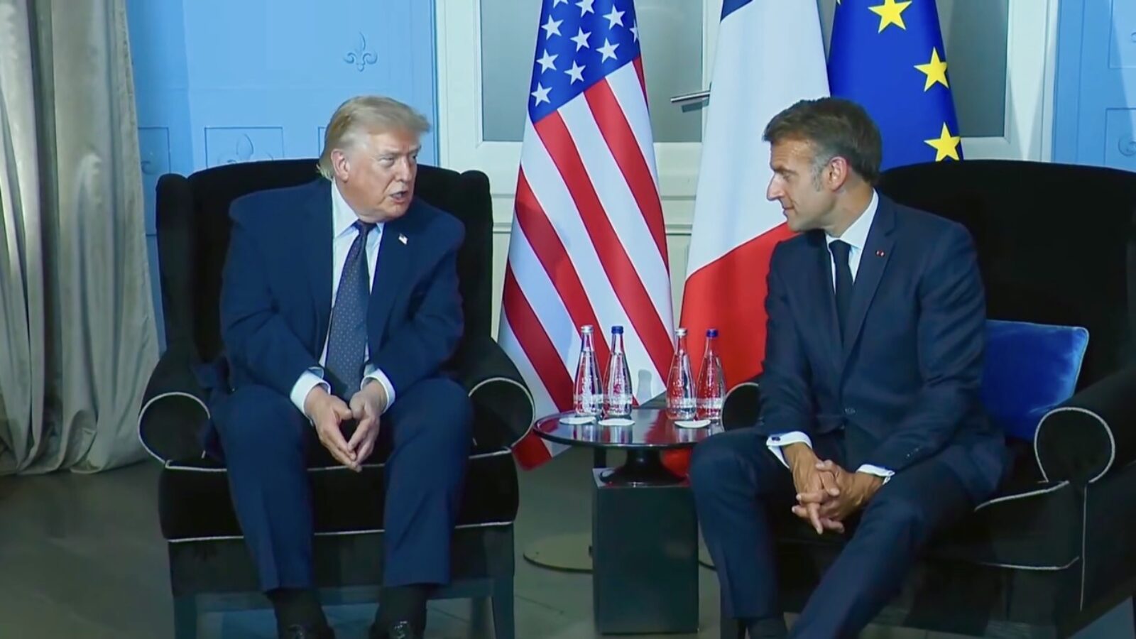 Donald Trump and Emmanuel Macron engage in a discussion during a diplomatic meeting, with flags of the United States and France visible in the background.