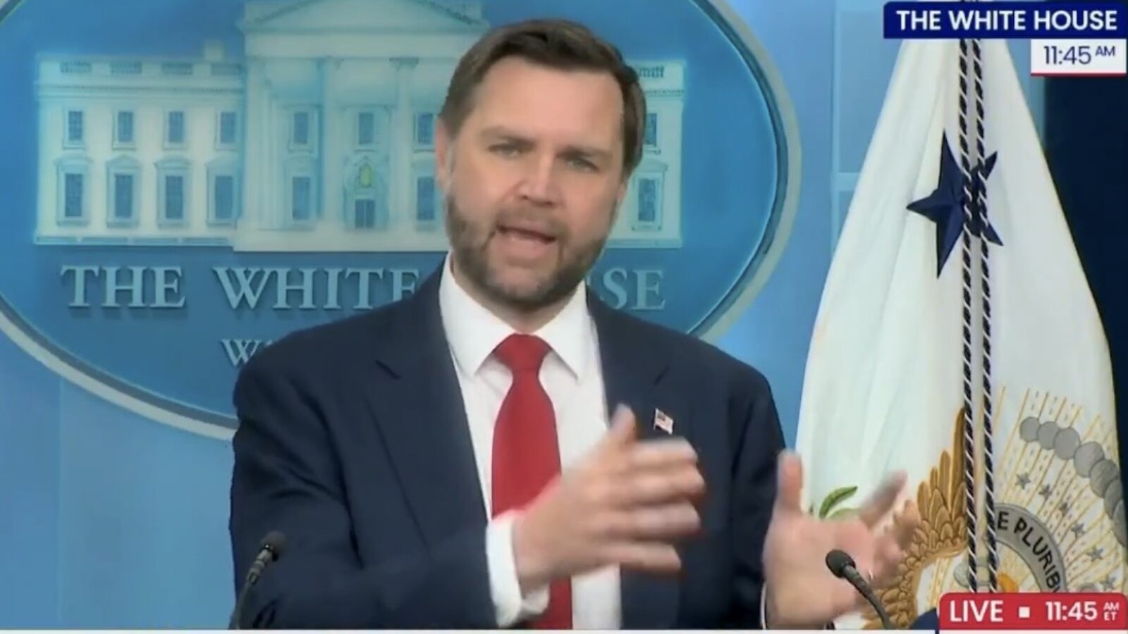 A man in a suit gestures while speaking at a press briefing in front of the White House backdrop.
