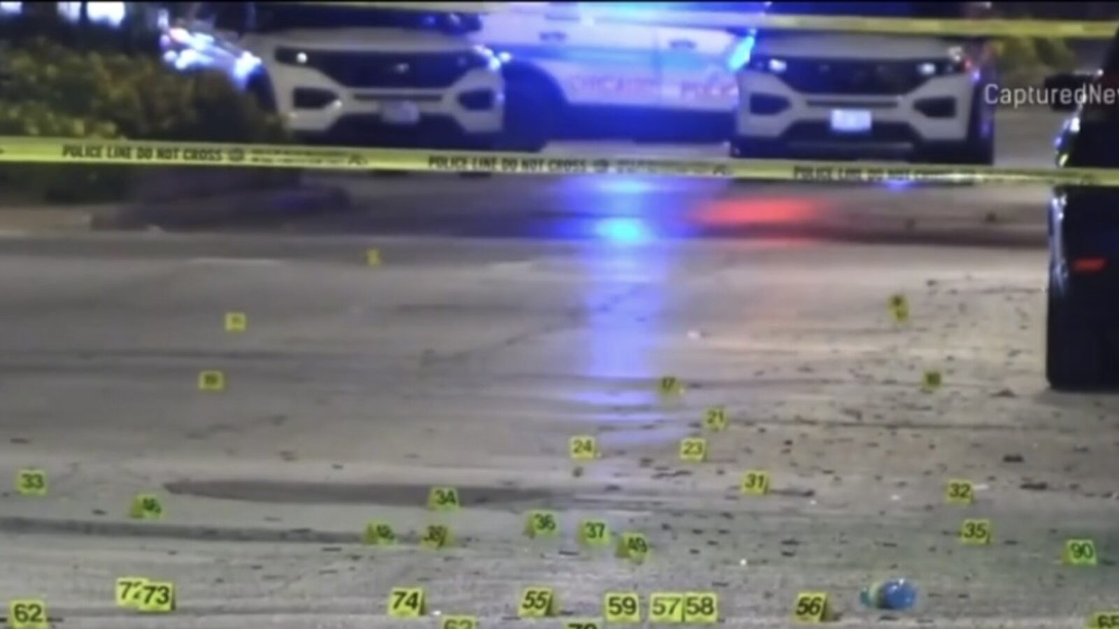 Police investigation scene with evidence markers on the ground, illuminated by police lights and surrounded by caution tape.