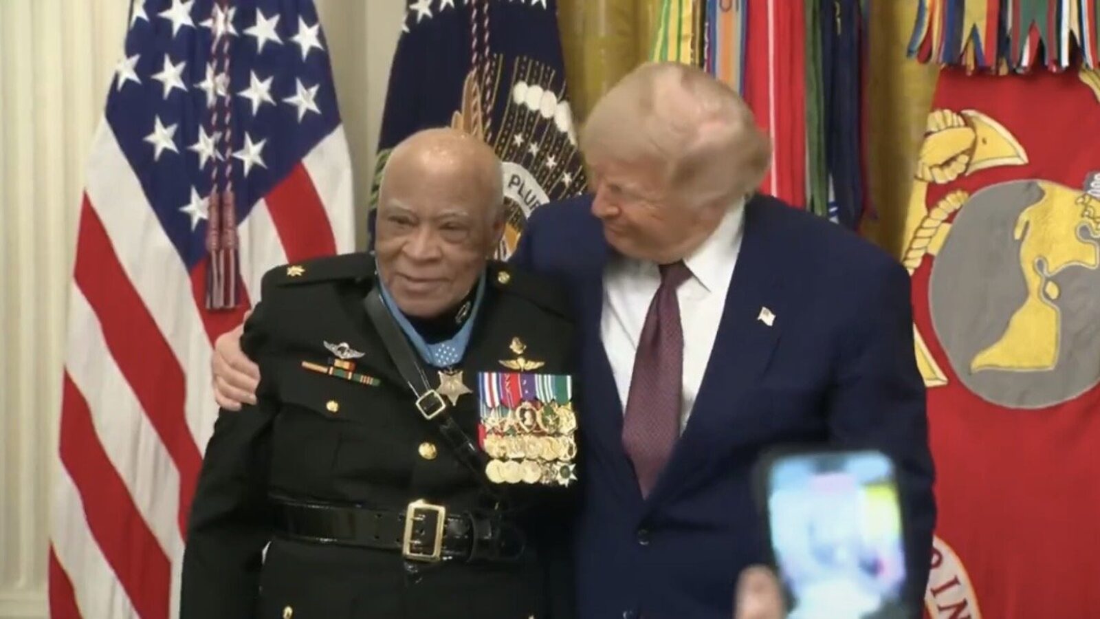 Donald Trump poses with a decorated military veteran, showcasing their camaraderie against a backdrop of flags and military honors.