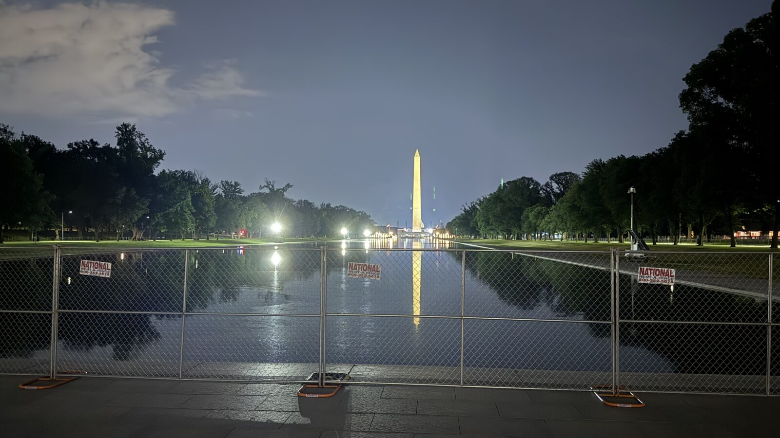 Washington Monument illuminated at night, reflecting in the water, with a security fence and trees in the foreground.