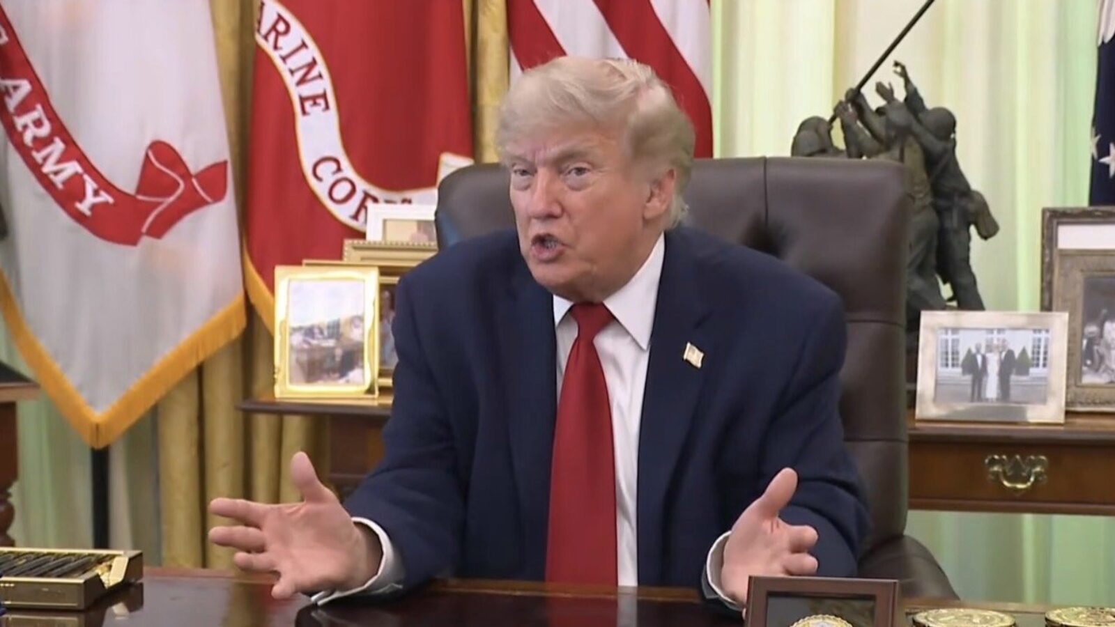 Donald Trump speaking passionately at his desk in the Oval Office, surrounded by U.S. flags and personal photos.