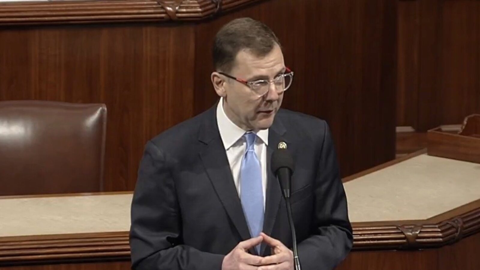 A man in a suit speaks at a podium in a legislative chamber, addressing an audience with a focused expression.