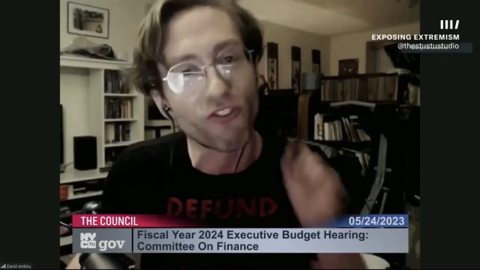 Person speaking during a virtual budget hearing, wearing a black shirt with the word "DEFUND," discussing financial matters in a home office setting with bookshelves in the background.