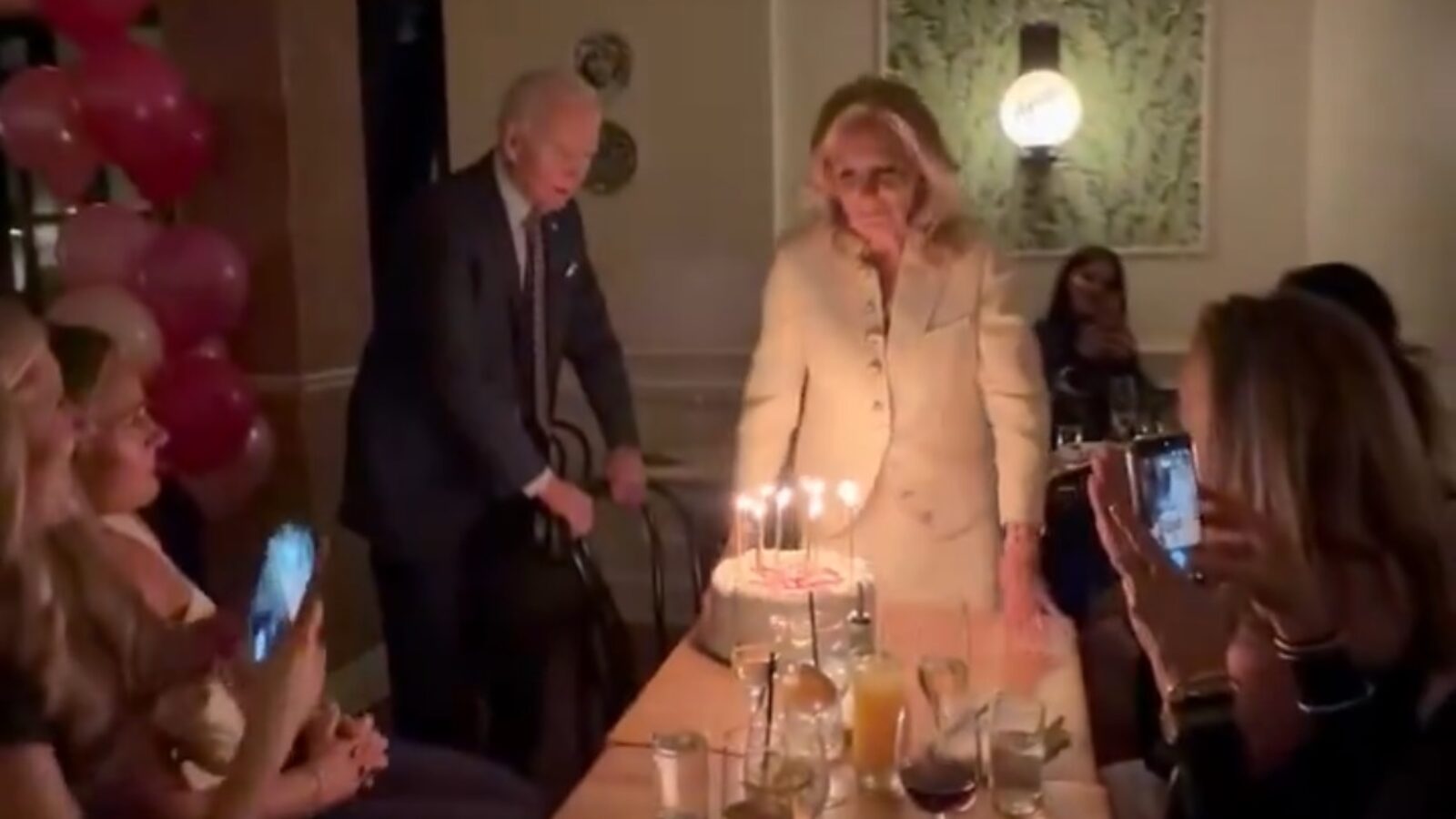 A birthday celebration featuring a woman approaching a cake with candles, surrounded by guests enjoying the moment and taking photos.