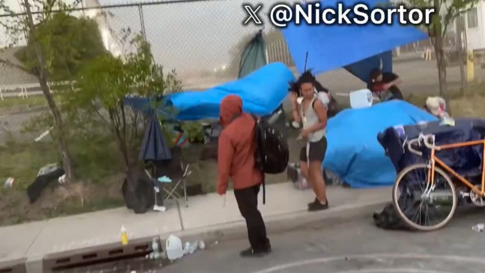 A group of individuals stands near makeshift shelters with blue tarps, highlighting homelessness and urban living conditions in an outdoor setting.