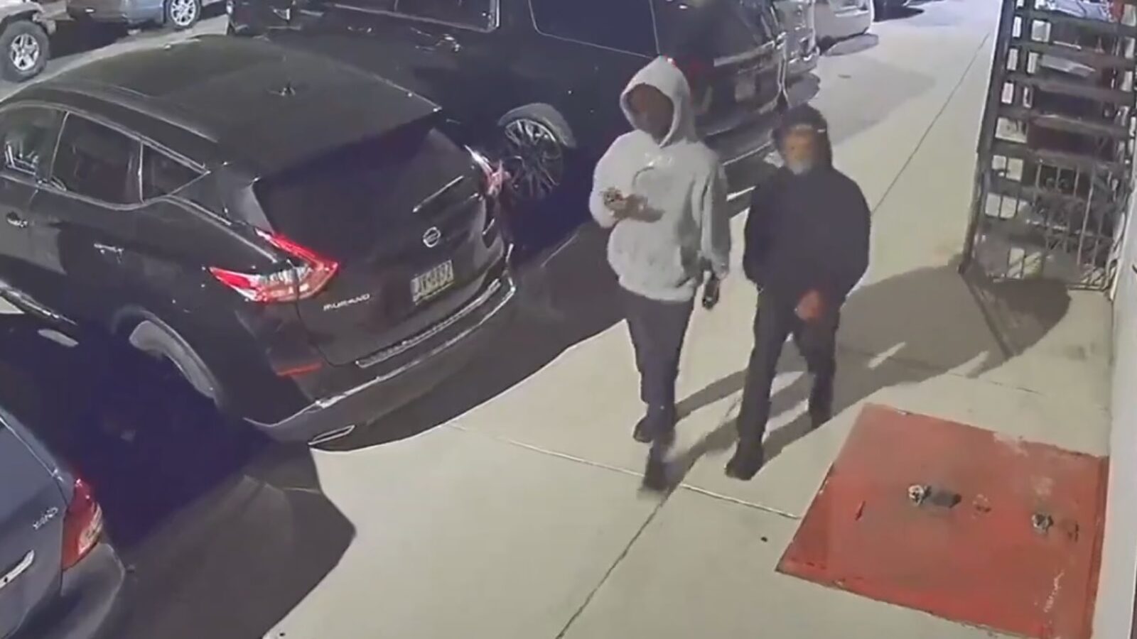 Two individuals walking past parked cars at night, one checking a phone, captured by security camera.