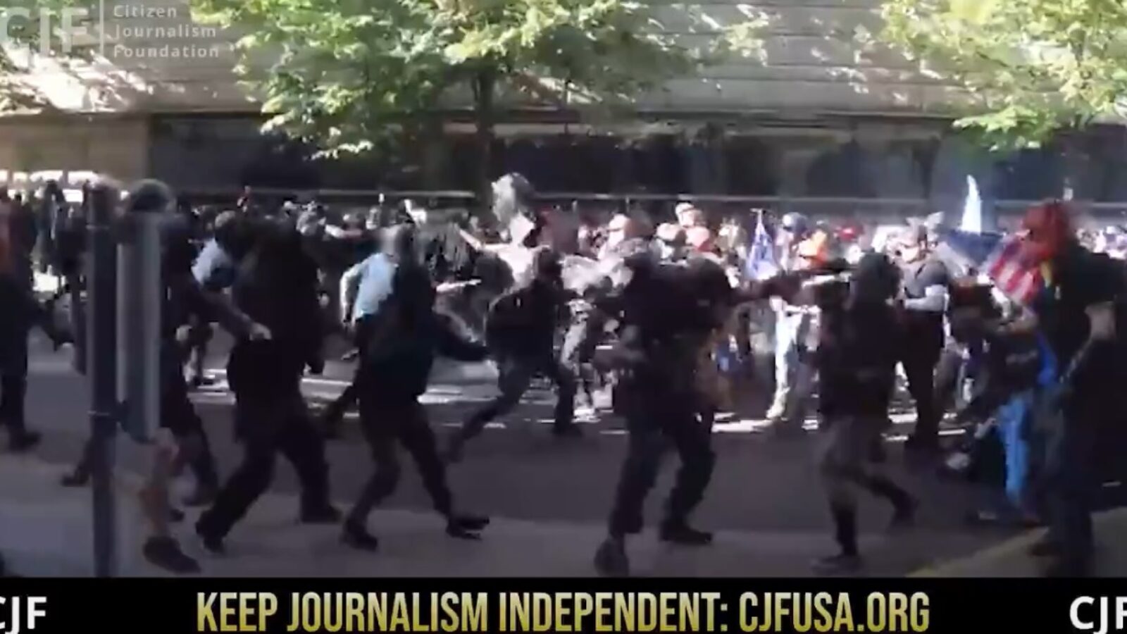 Protesters and law enforcement engage in a tense confrontation during a public demonstration, with a crowd visible in the background amidst urban surroundings.