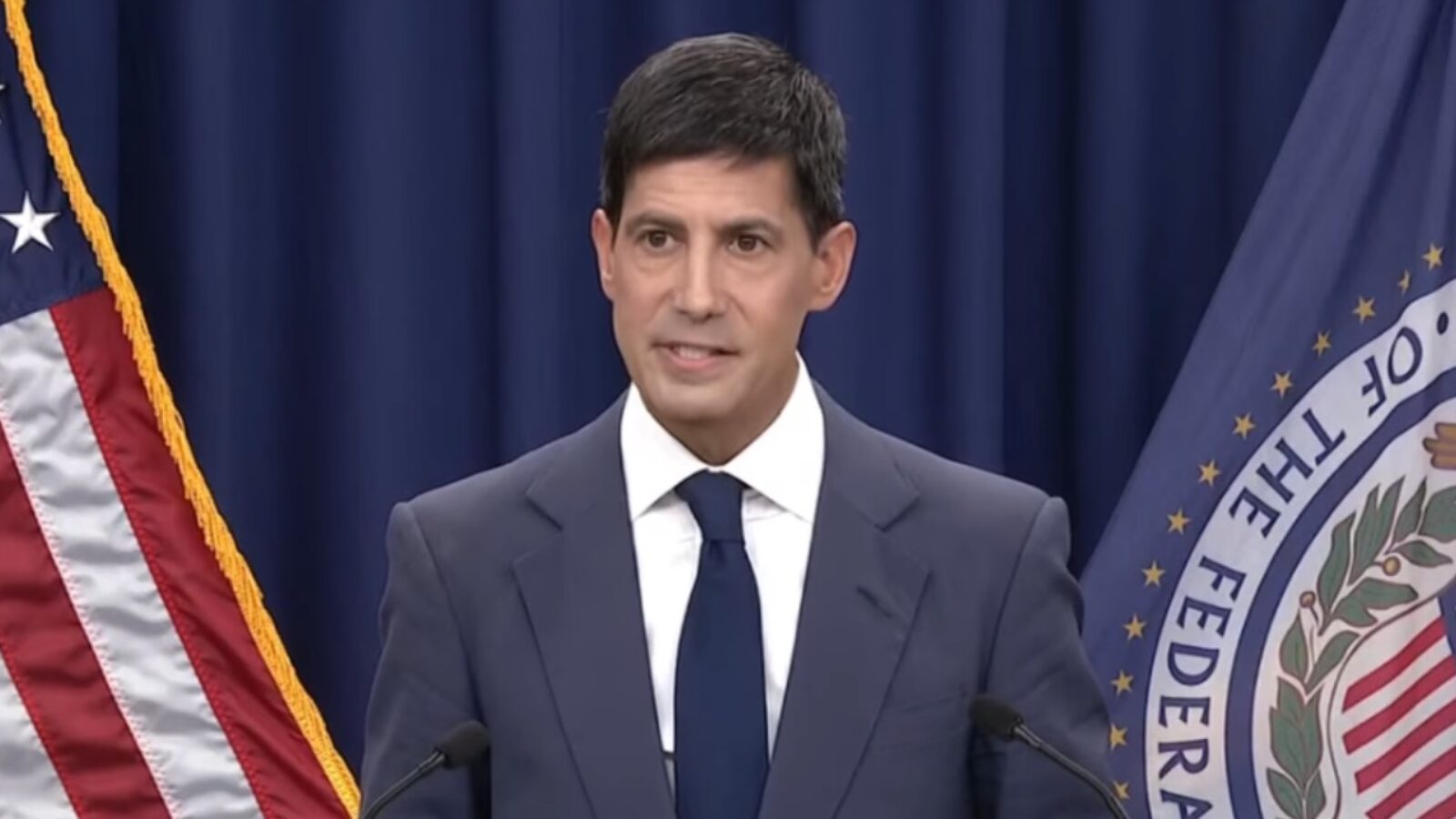 Official delivering a speech at a podium, flanked by the American flag and a state flag, emphasizing a significant announcement or policy.