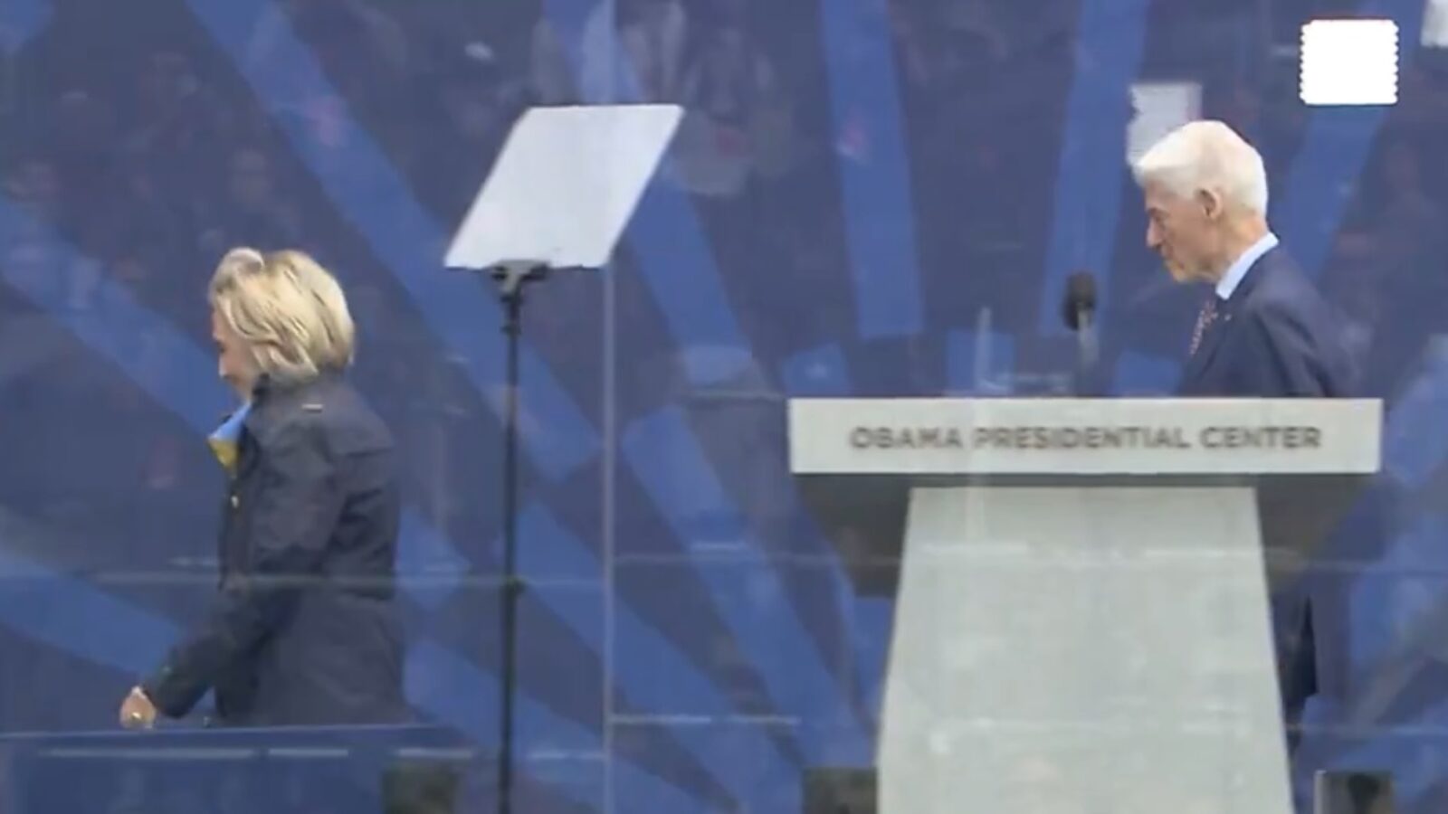 Hillary Clinton and Bill Clinton walking past the podium at the Obama Presidential Center event, with an audience visible in the background.