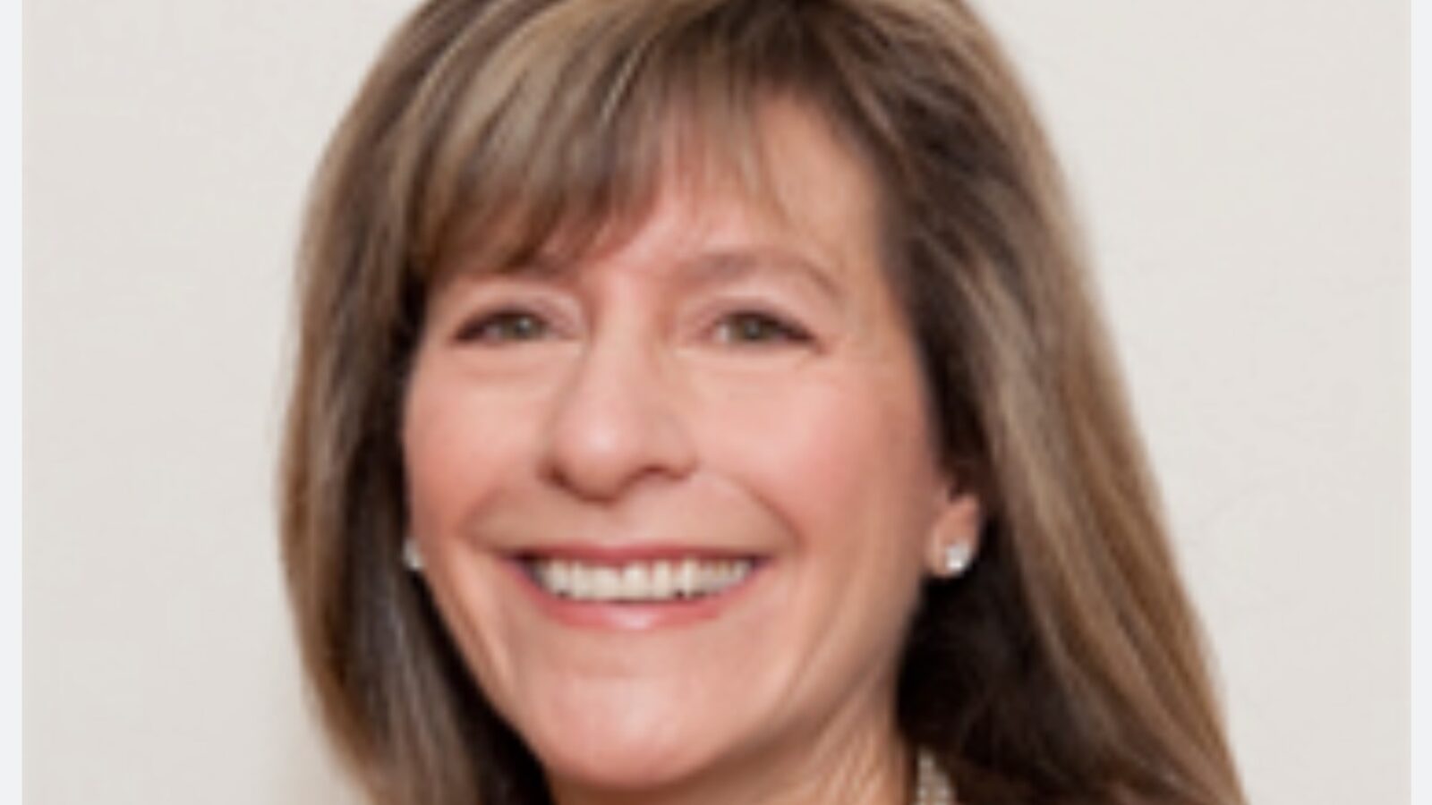 Smiling woman with shoulder-length brown hair, wearing earrings and a necklace, against a neutral background.