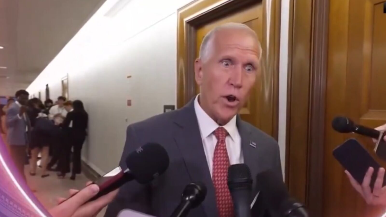 Politician speaking to reporters in a hallway, surrounded by media personnel and onlookers, highlighting a moment of public engagement.