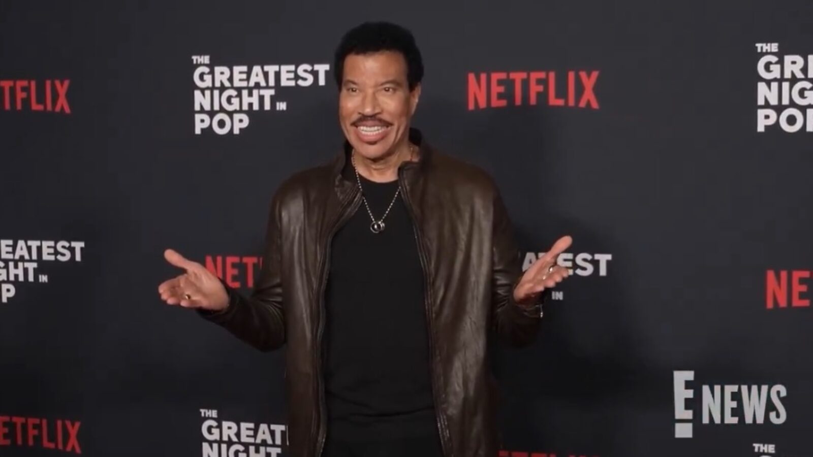 Lionel Richie smiles and gestures at the premiere event for The Greatest Night in Pop, hosted by Netflix.