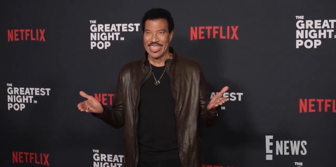 Lionel Richie smiles and gestures at the premiere event for The Greatest Night in Pop, hosted by Netflix.