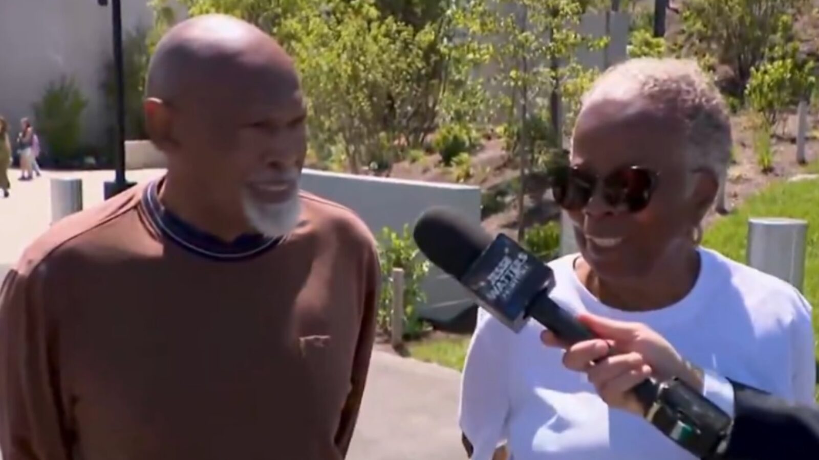 Elderly couple smiling while being interviewed outdoors, surrounded by greenery and people in the background.