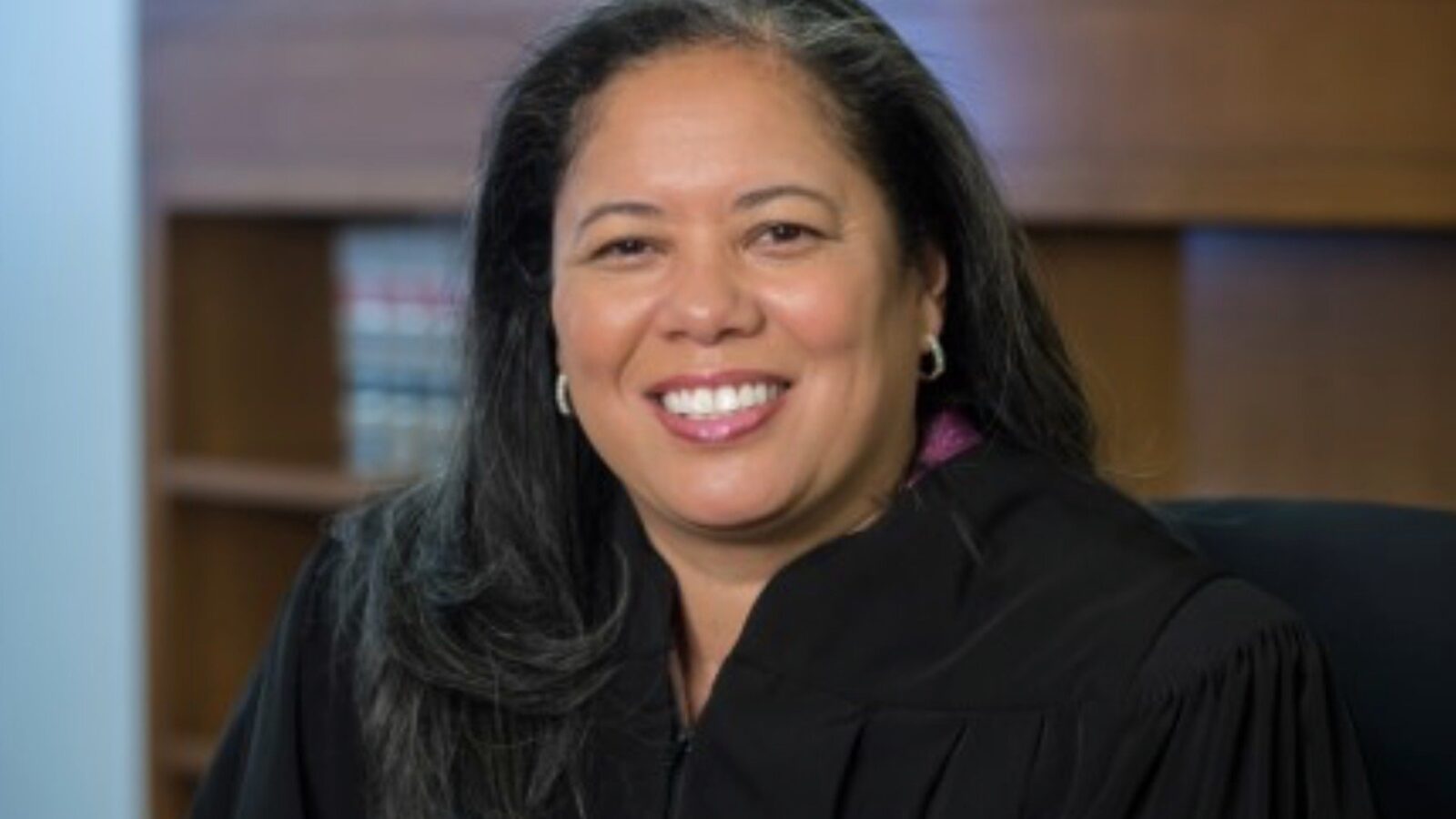 Smiling female judge in a black robe seated in an office with law books in the background, representing authority and professionalism in the legal field.