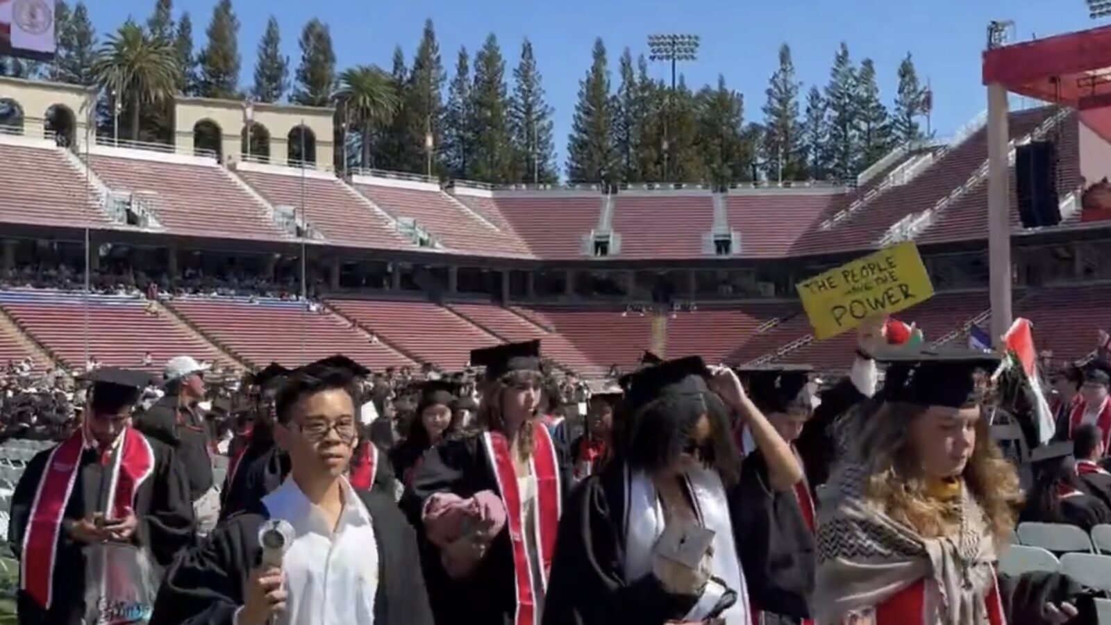 Graduates in caps and gowns walk through a stadium during a commencement ceremony, with one holding a sign that promotes empowerment.