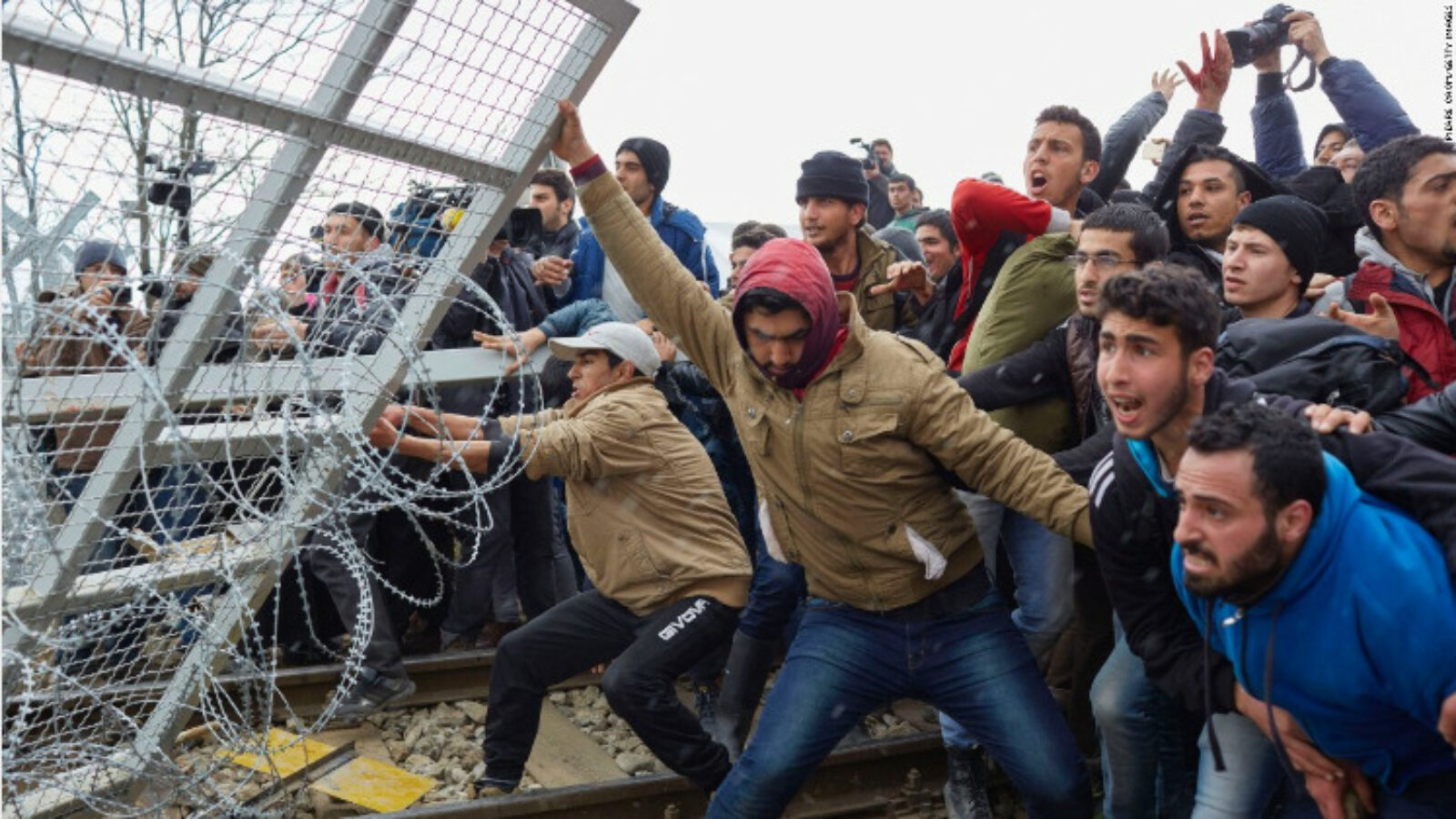 Group of young migrants attempting to breach a border fence amid a tense situation, showcasing the challenges of migration and border control.