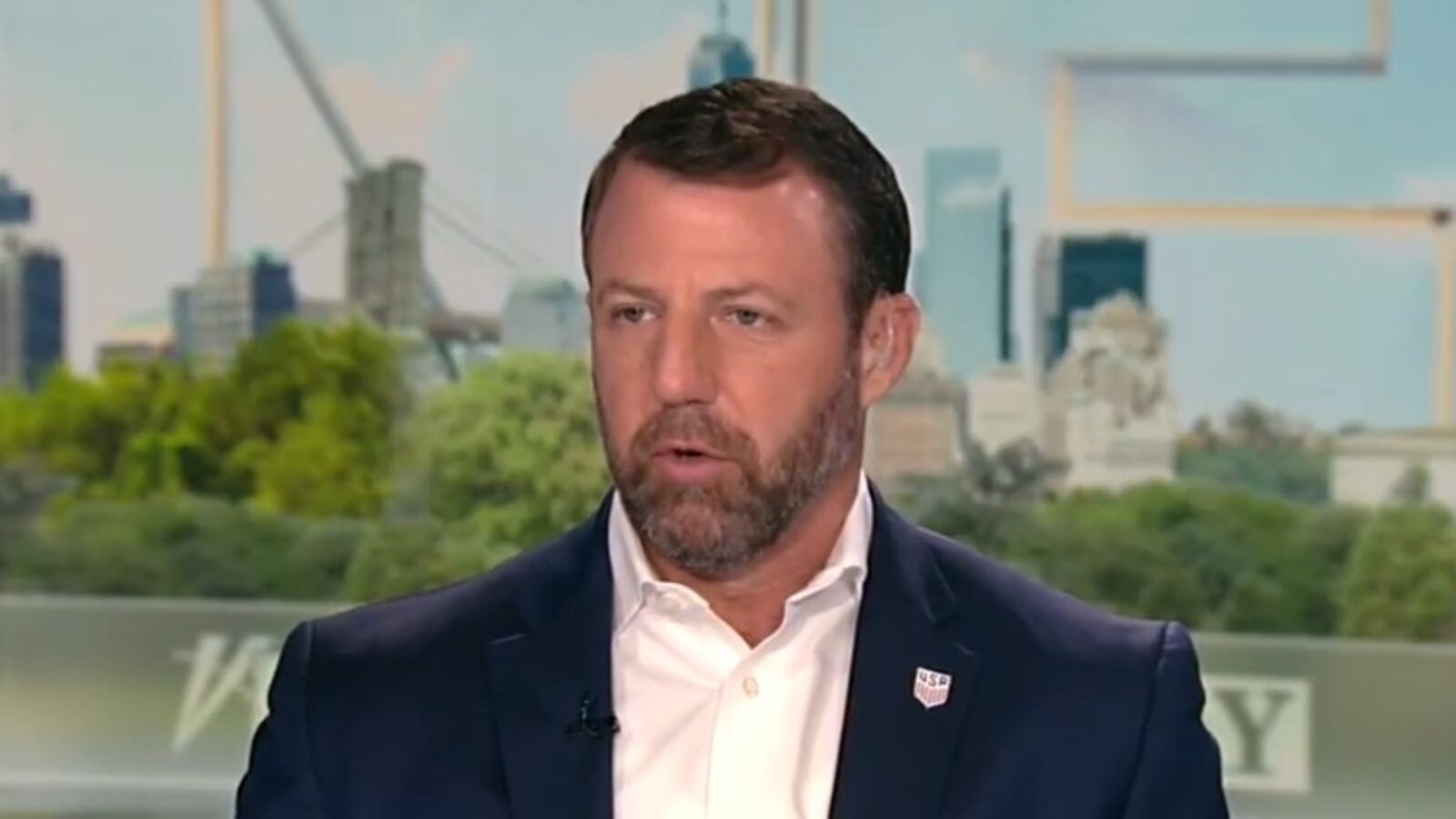 Man in a suit speaking during a television interview with a city skyline in the background, discussing current events and issues.