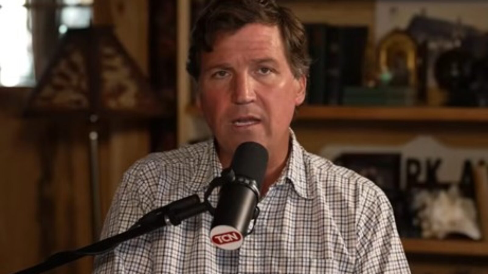 Tucker Carlson speaking into a microphone during a podcast recording in a cozy, rustic setting with books and decor in the background.