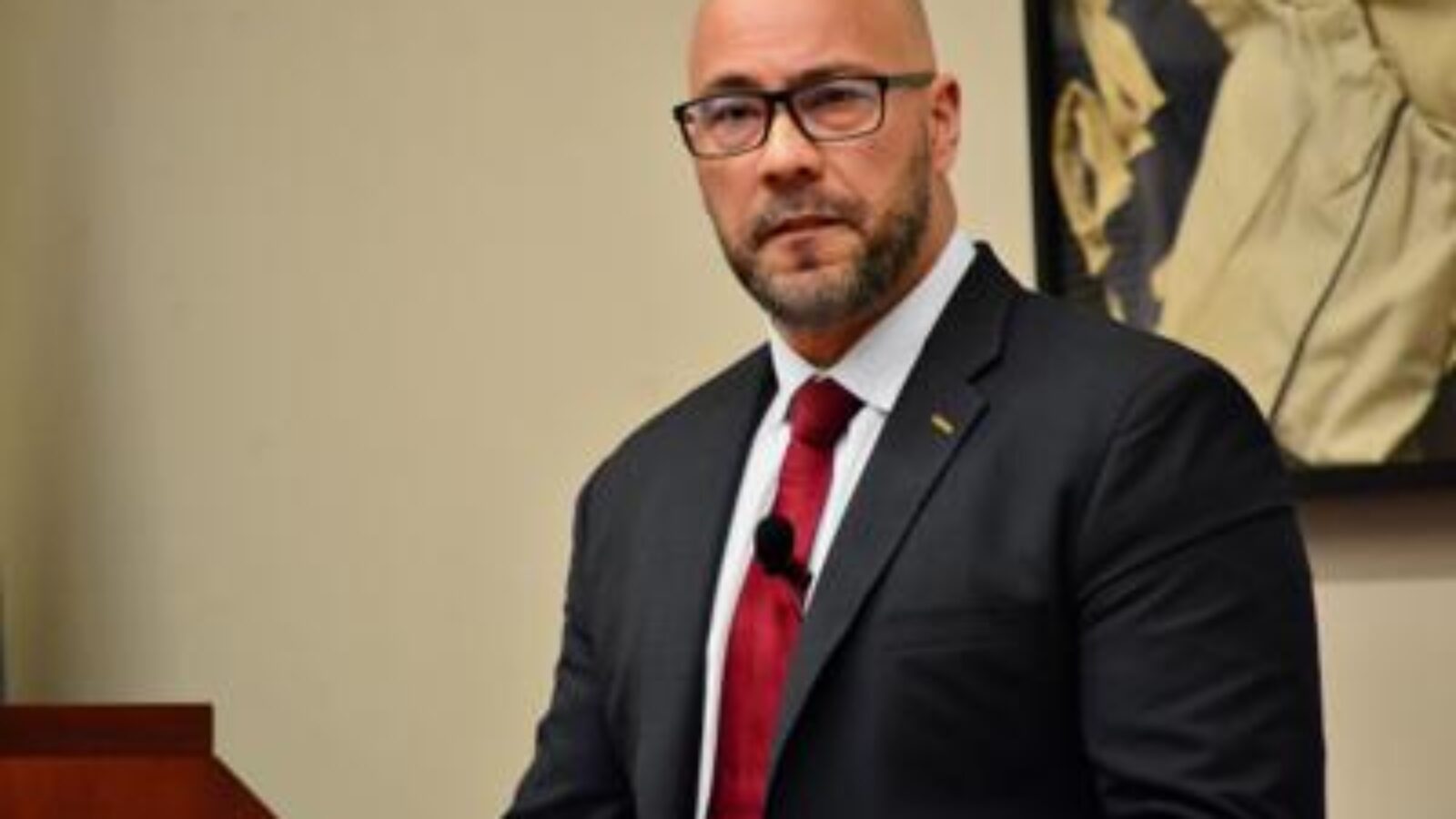 Business professional in a suit and glasses speaking at a podium during a conference or presentation.