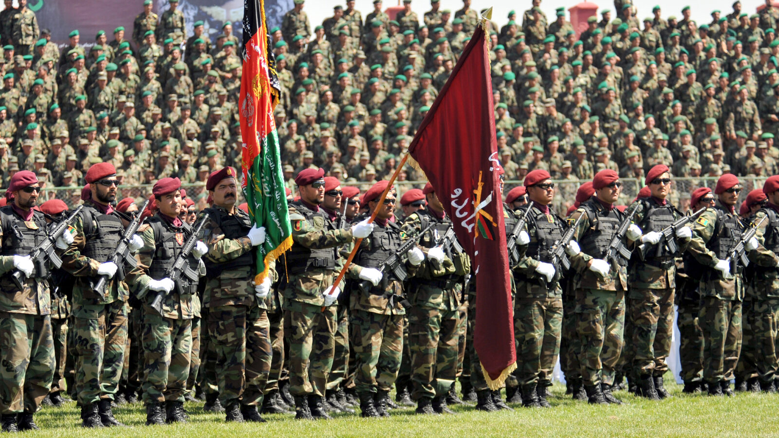 Afghan soldiers in military uniforms stand in formation during a parade, holding flags and weapons, with a large crowd of troops in the background.