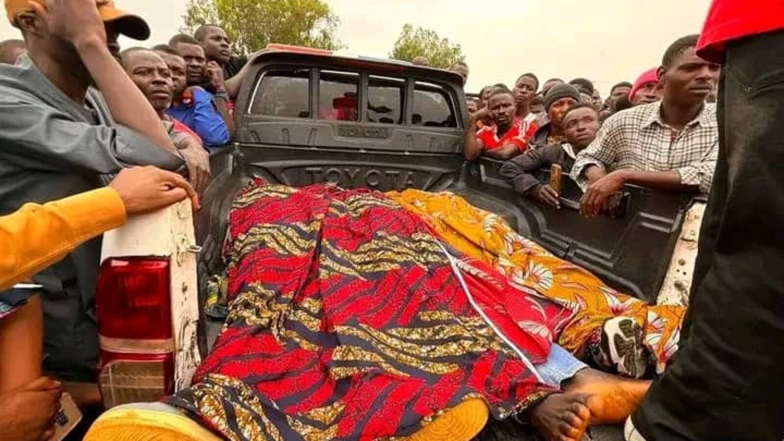 Crowd gathered around a pickup truck, observing a covered body, highlighting community reactions to a tragic event.