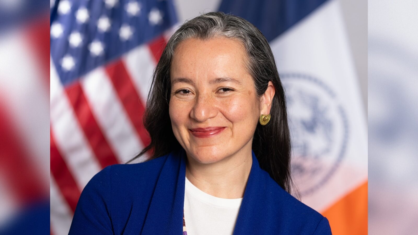 Portrait of a professional woman smiling, wearing a blue blazer, with American flags in the background, representing a government or public service context.
