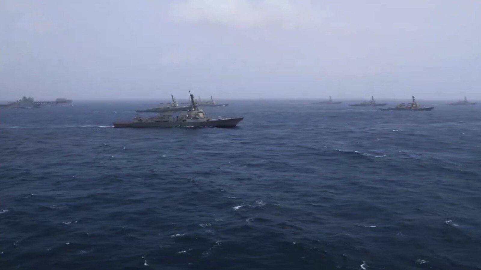 A fleet of military ships navigates through open waters under a cloudy sky, showcasing naval operations at sea.