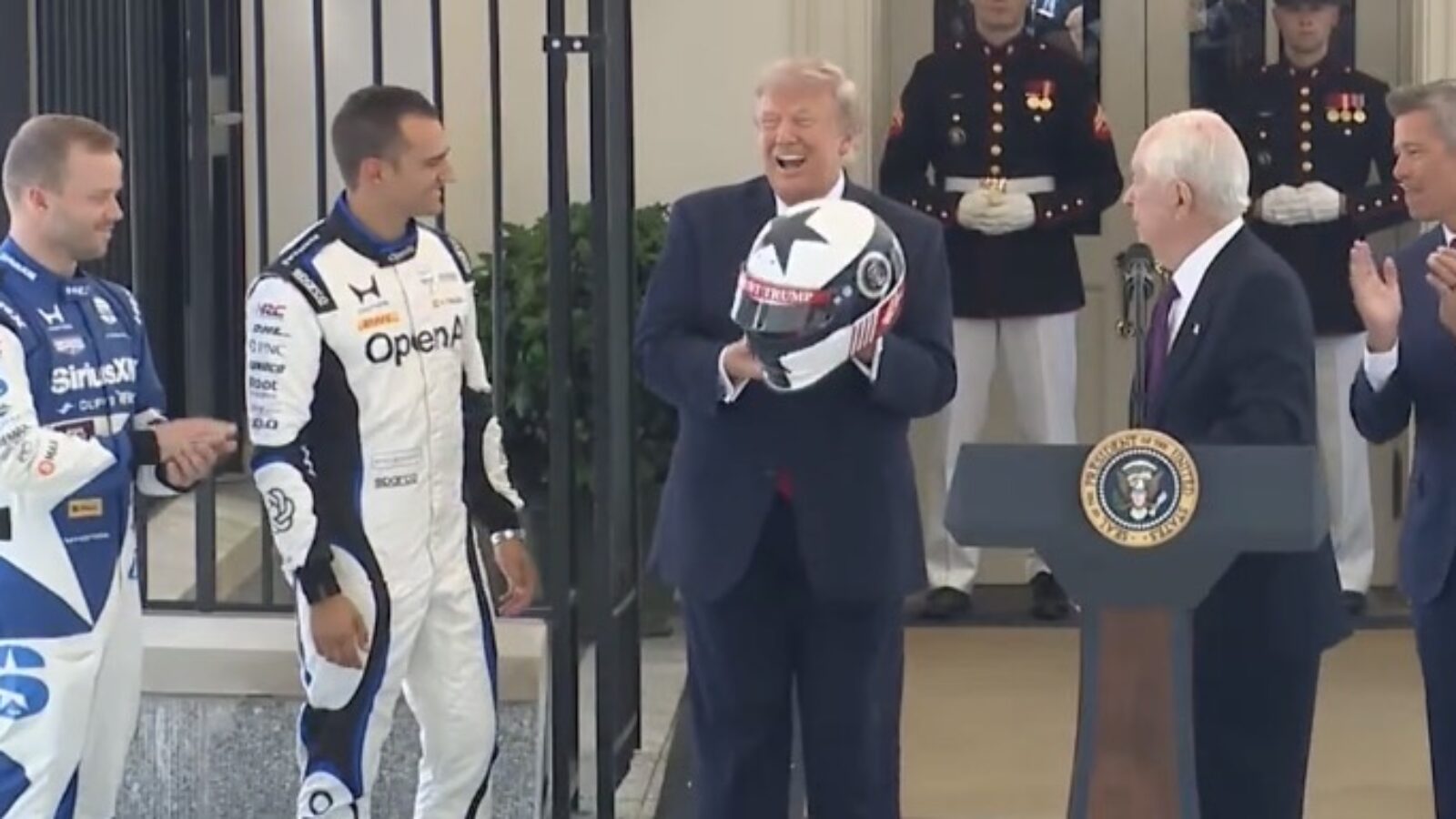 Donald Trump holds a racing helmet while smiling and interacting with race car drivers and officials during a ceremonial event at the White House.