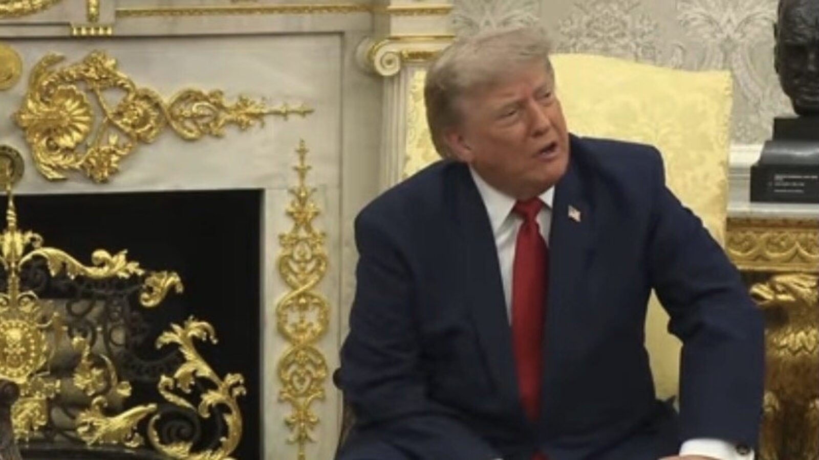 Donald Trump seated in a formal setting, engaging in conversation, with ornate decor and fireplace in the background.